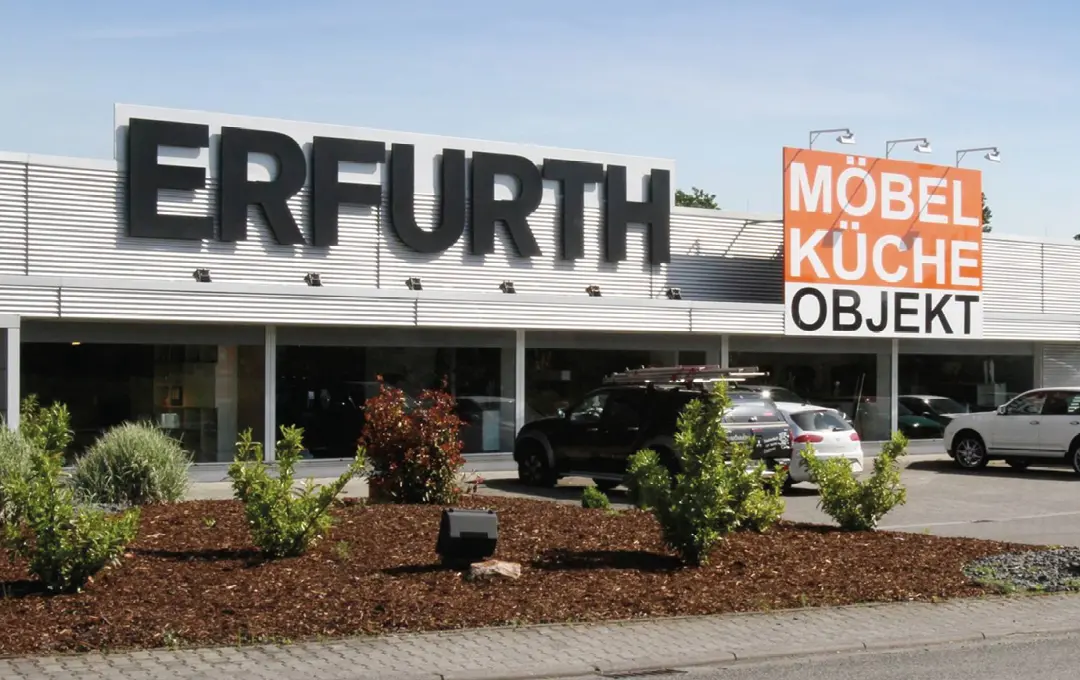 Außenansicht von Möbelhaus Erfurth mit großem Schriftzug und Schild Möbel Küche Objekt, davor Parkplatz, Sträucher und Straße.